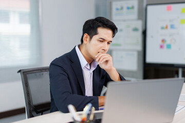 Asian businessman in suit is sitting at desk and thinking about solving a problem while working on...