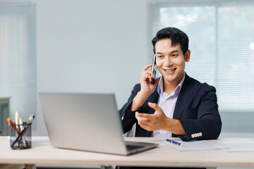 Young businessman smiling and gesturing while talking on smartphone and looking at laptop screen in...