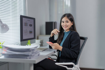 Young professional woman sitting at her desk using her smartphone and looking at camera, smiling,...