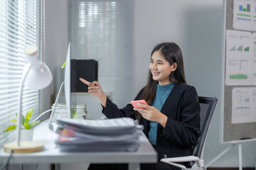 Asian businesswoman is sitting at her desk, pointing at her computer monitor while holding a...