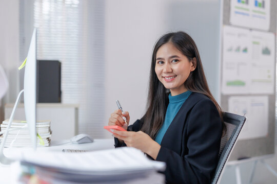 Young, happy Asian businesswoman smiling while taking notes on sticky notes, working efficiently at her desk in a modern office filled with technology and organized paperwork