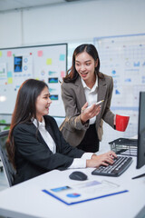 Two young businesswomen discussing work strategy in modern office, one is typing on keyboard while...