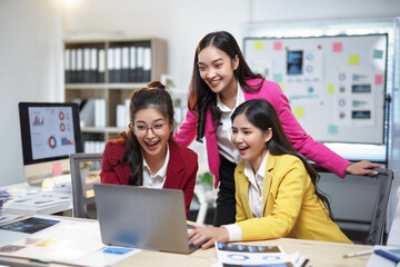 Three joyful businesswomen are looking at a laptop and celebrating a successful project in a modern...