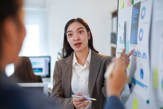 Asian businesswoman is presenting a project explaining charts on a whiteboard to colleagues during an office meeting, discussing strategies and analyzing financial data for business growth