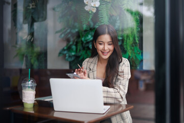 Smiling businesswoman using laptop and smartphone, managing her business remotely while enjoying a refreshing drink in a stylish cafe with green plants