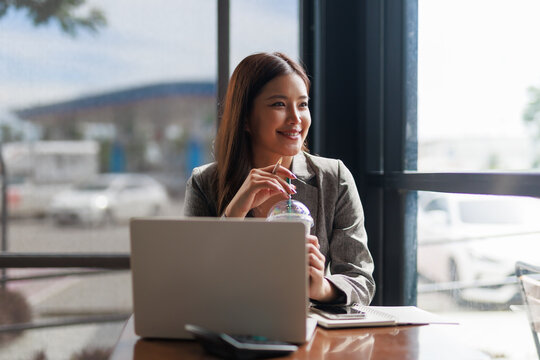Young asian businesswoman drinking a beverage and looking outside the window while working or studying on her laptop in a cafe, taking a break and enjoying the view - Powered by Adobe