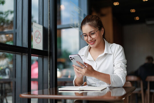 Smiling asian businesswoman using mobile phone while sitting at table in cafe with notebook near window with no smoking sign, enjoying break and using technology for work or communication
