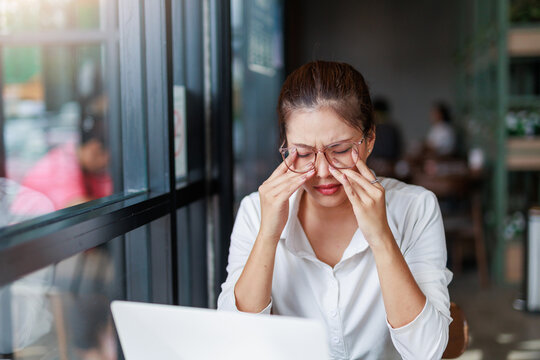 Young Asian businesswoman is working on laptop in cafe and feeling eye strain, rubbing tired dry irritated eyes, suffering from bad blurry vision after long laptop work - Powered by Adobe