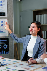 Asian businesswoman is holding a pen and pointing at charts and graphs on the wall while sitting at...