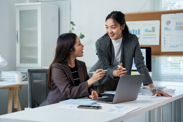 Two businesswomen engaged in a work discussion, collaborating on a project while using a laptop in a modern office environment. Sharing ideas and strategies enhances teamwork and communication