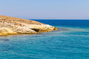 A tranquil scene featuring a crystal-clear turquoise sea gently lapping against a dry, rocky coastline under a cloudless blue sky. This peaceful landscape evokes the essence of summer and natural