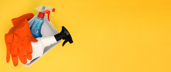 Spray bottles of cleaning agent and rubber gloves in white container on yellow background with place for text, top view