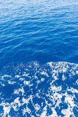 A vivid capture of the ocean's surface as a boat moves forward, creating swirling white foam patterns against the deep blue water. The texture of the sea and the energetic movement of the foam convey