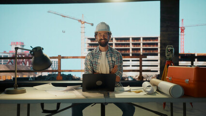 Confident architect in hardhat sits at his desk, overseeing construction project with cranes and building in background
