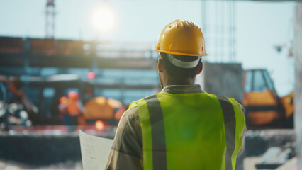 Construction manager supervising building site holding blueprints wearing safety vest and helmet