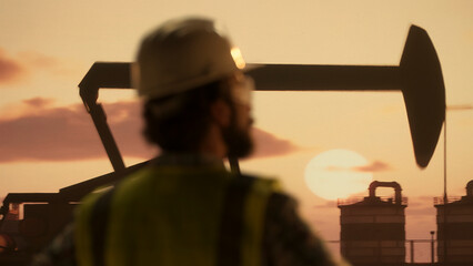 Oil industry worker watching pumpjack at sunset in oilfield