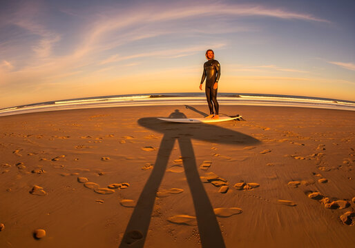 A surfer stands on their board on a sandy beach at sunset, casting a long shadow as the waves gently roll in.