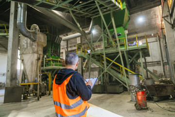 Supervisor writing notes on clipboard, inspecting wood processing machinery in a sawdust and wood...