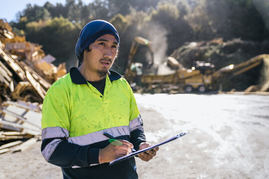 Waste management worker writing notes on clipboard while inspecting recycling operations at a landfill site, with heavy machinery working in the background - Powered by Adobe