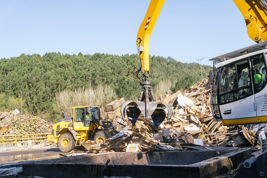 Claw of a material handler picking up wooden waste for recycling in a waste treatment plant, with a wheel loader and forest in the background