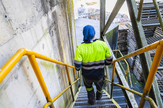 Factory worker dressed in high visibility clothing, climbing metal stairs within a recycling plant, focused on safety and efficiency - Powered by Adobe