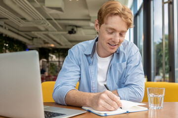 Ginger young man sitting at the table in the cafe and making notes