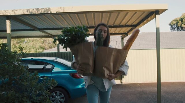 Young woman carries paper bags filled with groceries from blue car parked in residential driveway. Peaceful suburban setting during daytime creates calm atmosphere.
