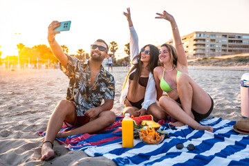 Friends taking a selfie on the beach at sunset while enjoying snacks and drinks