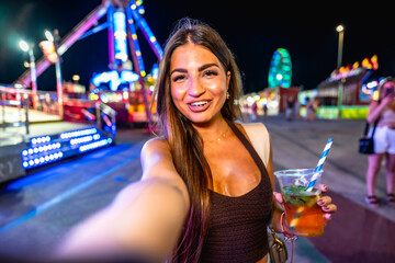 Young woman taking selfie at amusement park holding cocktail at night