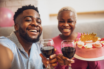 Happy young couple celebrating woman's birthday at home. Woman is holding cake and they are toasting with wine.