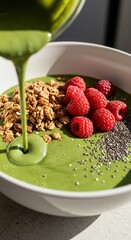 Vibrant Green Smoothie Bowl with Raspberries Granola and Chia Seeds Being Poured.