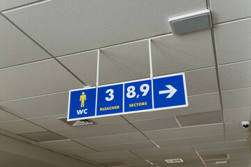 A blue sign with white arrows hanging on the ceiling in a public area indicating toilets, stands...