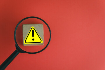 Hazard identification and safety concept. A magnifying glass inspecting a warning symbol on a block, representing a workplace risk or a product safety issue. OHS and caution alert.