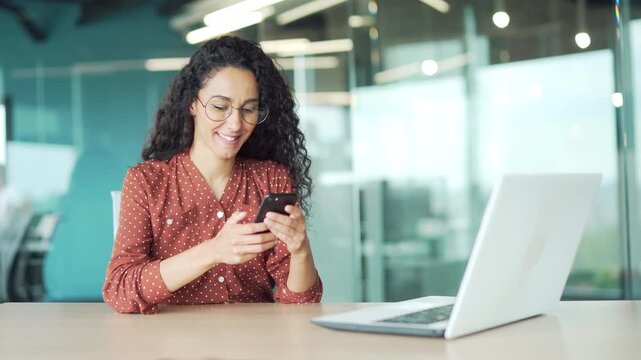Smiling young businesswoman using mobile phone sitting at workplace in modern business office. Happy female employee browsing web, chatting online, typing, reading message, banking or checking email