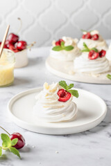 Meringues mini pavlova cake with whipped cream, fresh cherry and mint leaves on a plate, white marble background. Light summer dessert, copy space