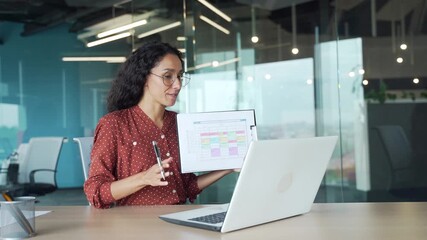 Young businesswoman speaks online by video call conference presentation using a laptop while sitting at workplace in a business office. Confident female manager communicates remotely, the graph shows - Powered by Adobe