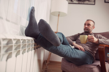 Man with tea cup holding feet with socks on a home heating radiator on a cold winter day outdoors.