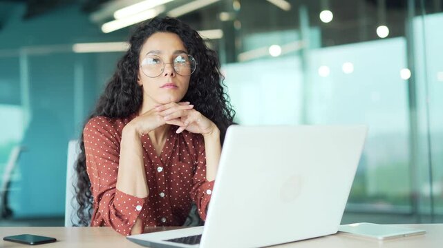 Busy businesswoman thinking about the task while working on laptop sitting at workplace in a modern business office. Thoughtful young female employee deals with project, thinks about solving a problem