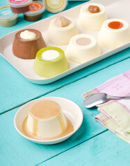 An assortment of colorful and creamy panna cottas or puddings with various toppings are presented on a white tray and a separate dish, arranged on a vibrant turquoise wooden surface