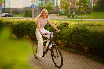 woman cycling through city park enjoys sunny day. dressed in casual white attire, rides bike along paved path lined with lush greenery. urban lifestyle, nature harmony
