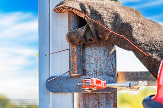 A close-up view of a gloved worker's hand securing two rusted metal beams with a clamp during an outdoor welding or metalworking operation.