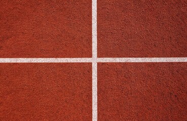 Top view of red running track surface with white lines. Geometric pattern of intersecting lines on textured ground. High angle shot emphasizes symmetry and stadium court.