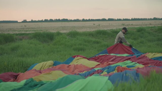 man in cap and gloves sets out colorful hot air balloon canopy across grassy field at dusk under pastel sky during preflight setup in expansive rural landscape