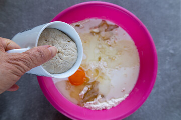 A hand pours a cup of activated yeast into a pink mixing bowl filled with flour, milk, an egg, and other ingredients. 