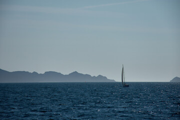 A sailboat sails at sunset through the Vigo Estuary, with the silhouettes of the Cies Islands outlined on the horizon and the shimmering waters.