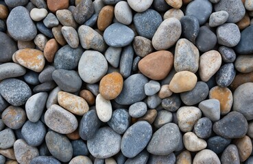 Collection of various sized rocks, predominantly gray, brown, orange, and yellow. Smooth, tumbled stones create a textured surface. Natural background suggests a beach or riverbed setting.