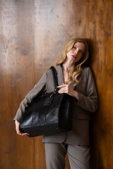 A stylish young woman, long blonde hair, poses in a grey suit, holding a black leather bag against...