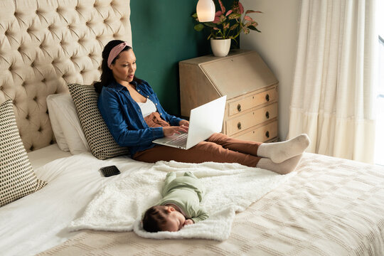Mother and baby sitting on tufted bed among pillows in bedroom typing on laptop, copy space