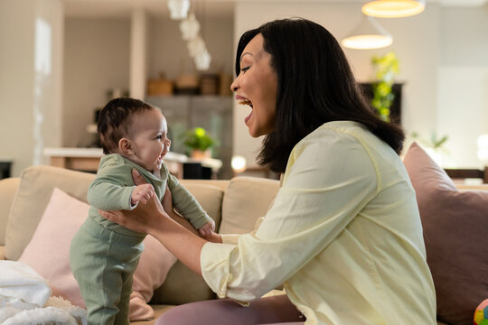 Asian mother and daughter giggling while holding baby on beige sofa at home with pendant lights