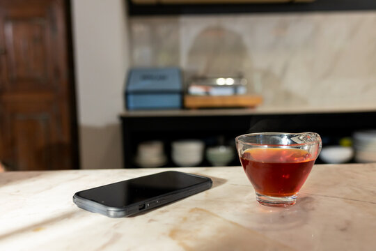 Smartphone lying beside cup of dark tea on marble countertop, with blurry bowls and boxes behind